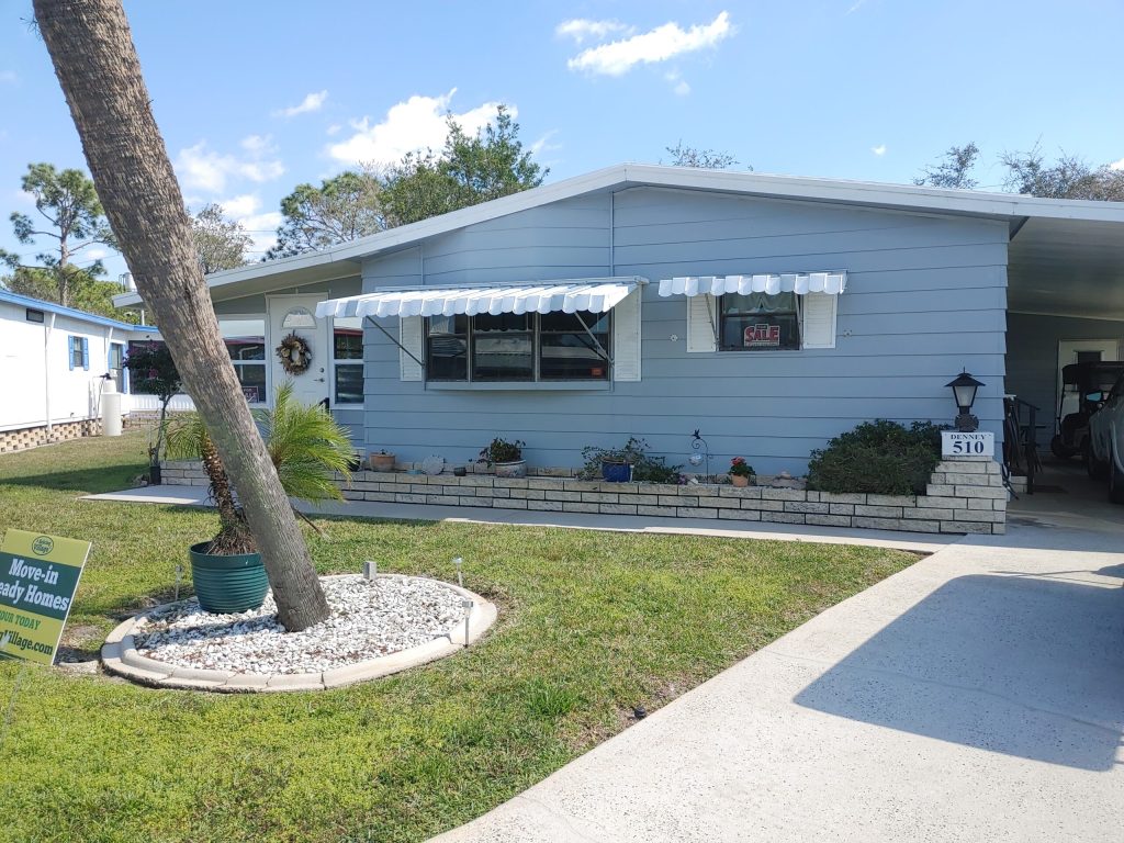 Single-story manufactured home with light blue siding, white trim, and awnings. A "For Sale" sign is visible in the window. There is a driveway, small garden, and a yard sign.