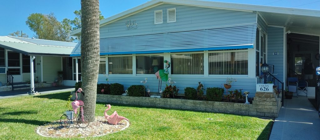 A blue single-story house with a covered porch, manicured lawn, garden decorations, and address sign reading "626" by the driveway.