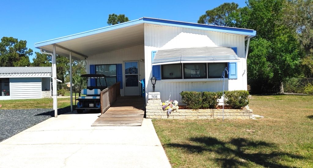 A white manufactured home with blue trim, a covered carport, wheelchair ramp, front steps, and a small landscaped yard with bushes and flowers.