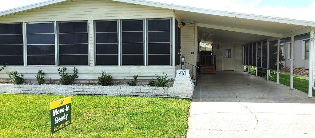Single-story manufactured home with large screened windows, attached covered carport, concrete driveway, and a "Move-in Ready" sign posted on the front lawn.