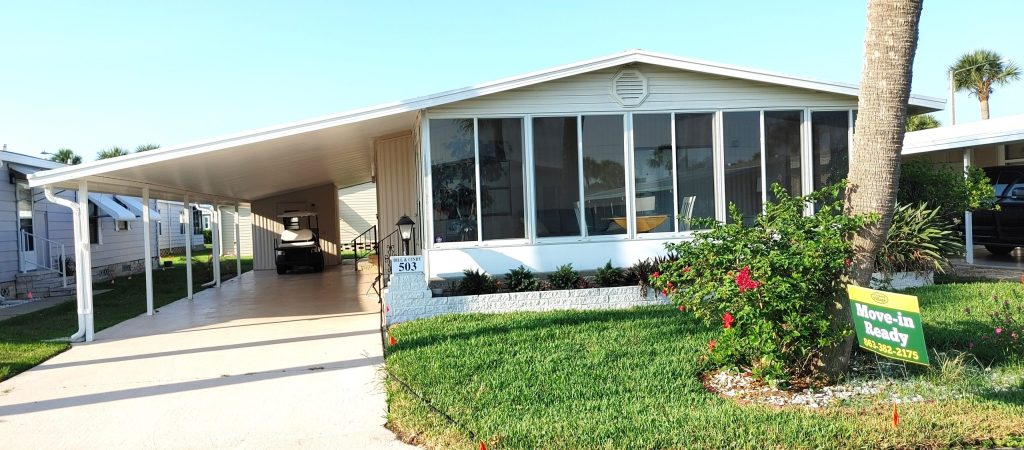 Single-story manufactured home with a covered carport, screened front porch, and a "Move-in Ready" sign on the front lawn surrounded by green grass and landscaping.