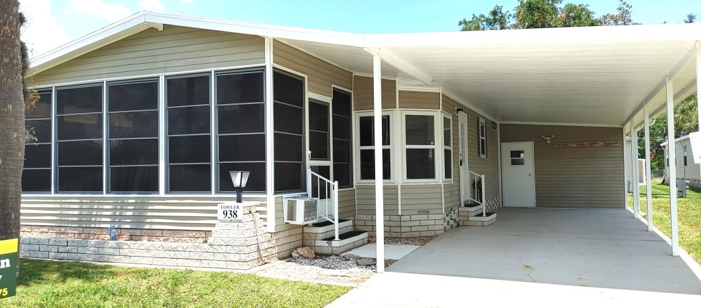 Single-story manufactured home with screened porch, covered carport, and small steps leading to the front door, set on a concrete driveway.
