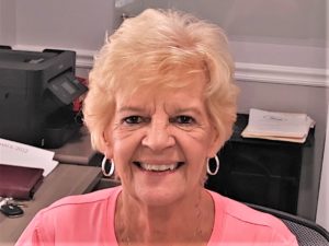 An older woman with short, curly blonde hair and hoop earrings smiles at the camera while sitting at a desk in an office setting.
