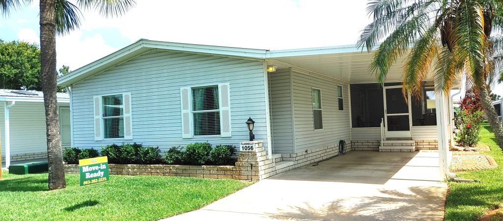 White manufactured home with a carport, palm tree, landscaped yard, and a "Move-In Ready" real estate sign in the front lawn.