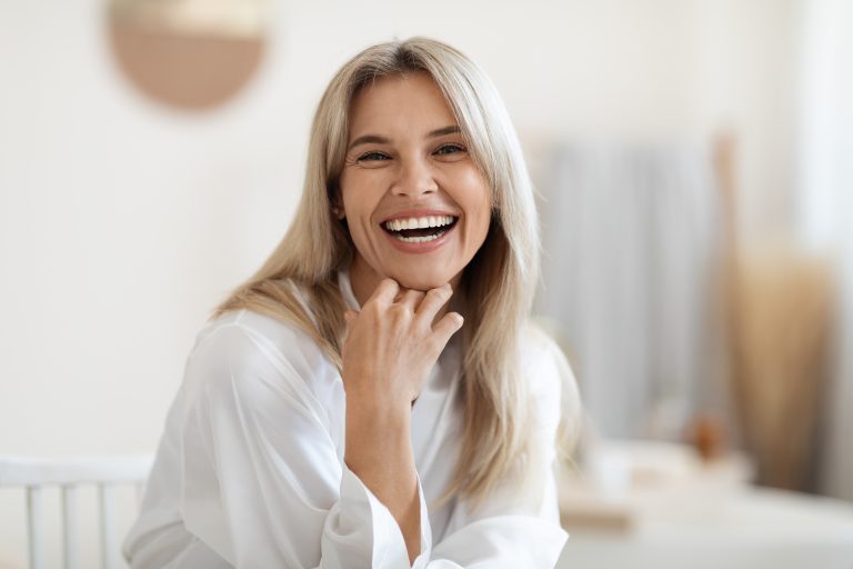 A woman with long blonde hair wearing a white blouse smiles and rests her chin on her hand in a bright, softly blurred indoor setting.