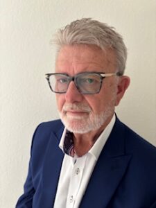 Older man with gray hair, beard, and glasses wearing a navy suit jacket and white shirt, standing against a plain light-colored wall.