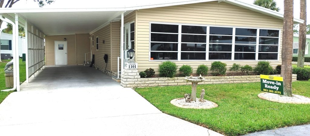 Single-story beige manufactured home with carport, large windows, landscaped front yard, and a "Move-in Ready" sign on the lawn.