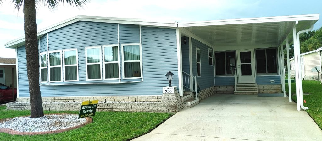 A light blue manufactured home with a covered carport, multiple front windows, and a "Move-In Ready" sign on the lawn. House number 926 is visible near the entrance.